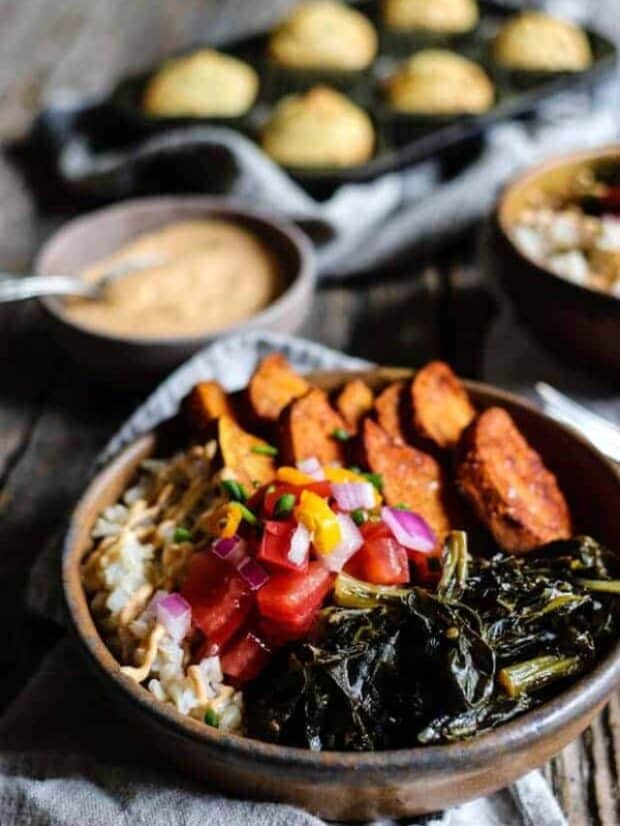 A bowl filled with brown rice, roasted sweet potatoes, collard greens, and pico de gallo sits on top of a line napkin on a table top. There is a small bowl of chipotle cashew cream in the background as well as pan of cornbread muffins.