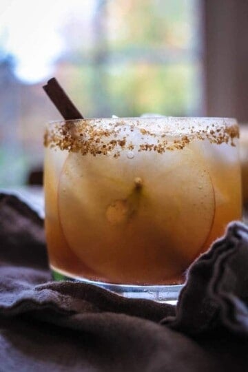 A cocktail glass filled with a gold colored cocktail, a very tin apple slice, and a cinnamon stick is sitting in front of a window that has trees with fall foliage outside of it. The glass has a brown sugar and cinnamon rim.