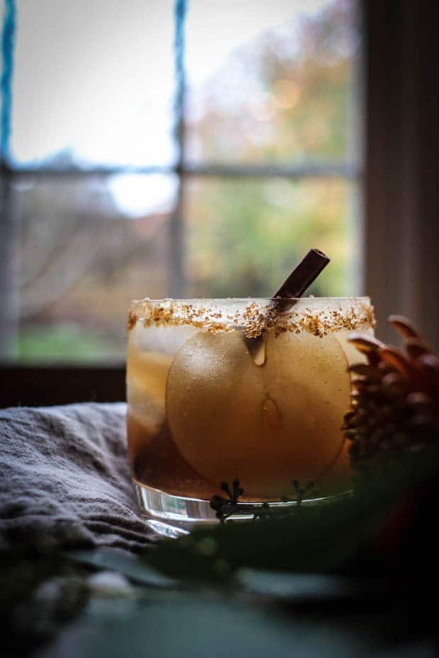 A festive cocktail with a thin slice of apple, a cinnamon stick, and a brown sugar cinnamon rim sits on a table with greenery and linens in front of a window that looks out upon trees that are full of autumn colored leaves. 