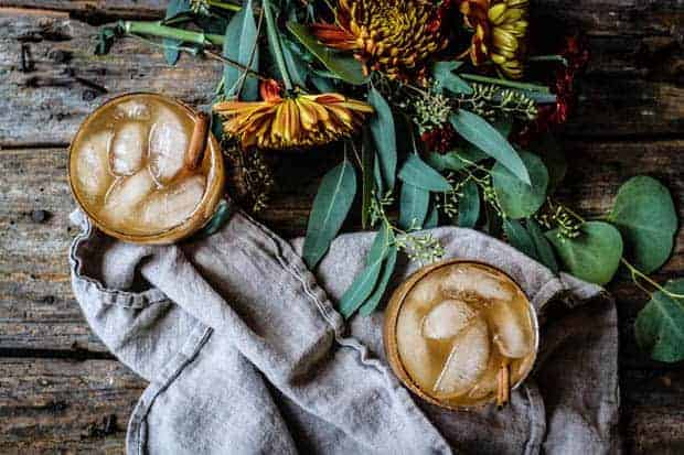 2 Spiked Apple Cider Cocktails with ice and a linen napkin are set upon a worn wooden table. There are also yellow and orange flowers laid on the table. 