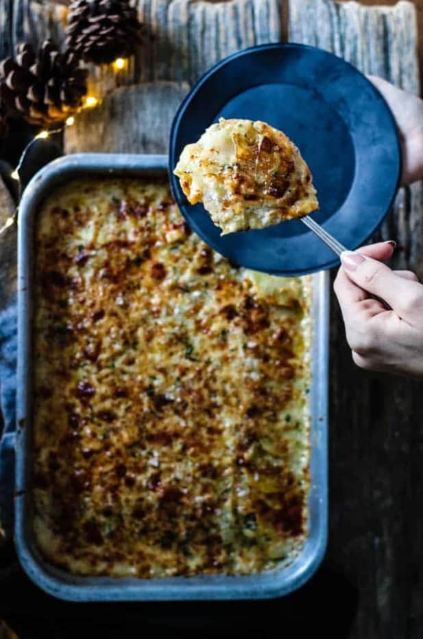 A baking pan filled with scalloped potatoes is sitting on top of a worn wooden table top. There is a linen napkin under the pan. Next to the pan on the table is a few pinecones and a small string of Christmas lights. A woman is scooping the scalloped potatoes onto a black plate.
