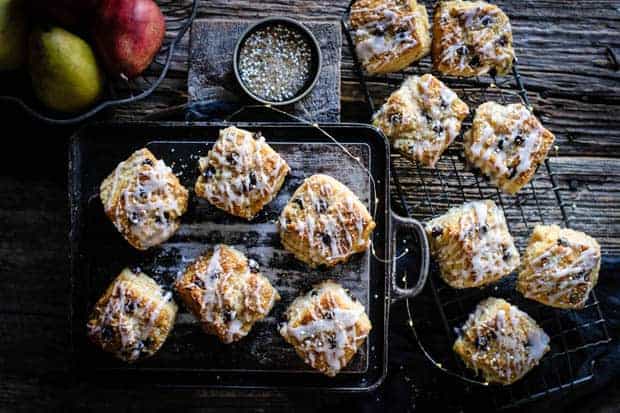 Dark chocolate pear scones that are drizzled with pear icing and sparkling sugar are sitting on top of a metal baking pan and some on a wire cooling rack. There are red and green pears in a bowl off to the side and a small black shallow dish filled with sparkling sugar on the table.