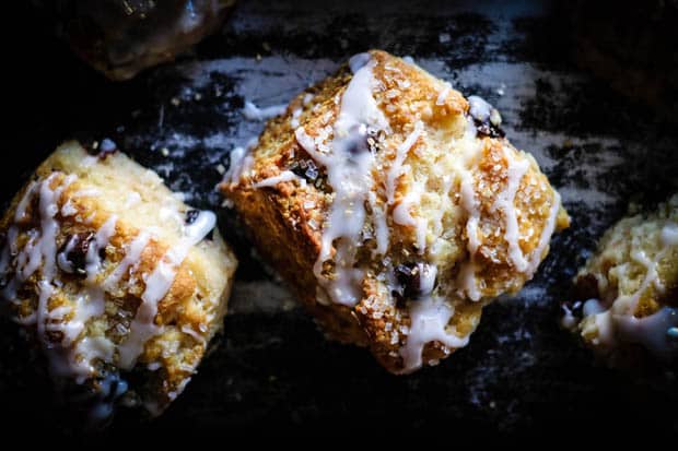 A close up image of a pear scone with dark chocolate chips, sparkling sugar, and fresh pear icing. The background behind the scone is black and silver metal.