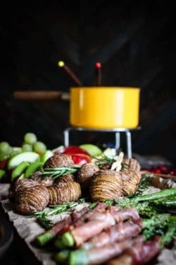 A vintage yellow fondue pot is in the background of the image on a wood board covered in brown parchment paper in front of the fondue pot is roasted prosciutto wrapped asparagus spears, hasselback potatoes, and sliced red and green apples.