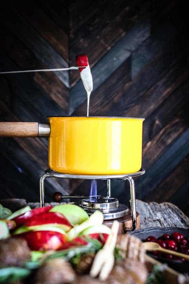 A slice of red apple is on a fondue fork. The fondue fork has dunked down in a yellow fondue pot and teh apple slice is half dipped in cheese fondue. In front of the fondue pot there is a pile of red and green apple slices, hasselback potatoes, and a bowl of pickled cranberries.
