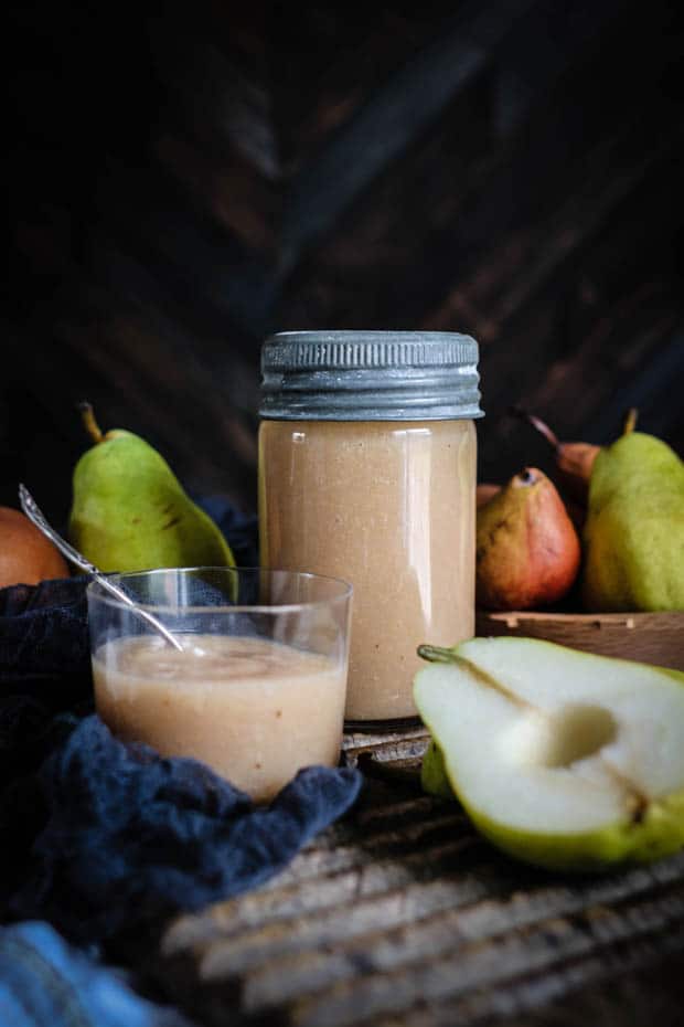 A glass jar filled with roasted pear sauce sits next to a glass cup of roasted pear sauce on a wooden table. There are green and reddish brown pears in teh background of the image and a sliced green pear with teh core removed in the front of the image. 