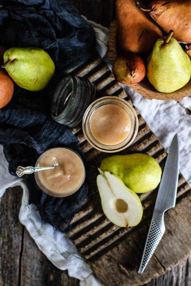 A glass storage jar and a small glass cup are filled with roasted pear sauce. the cup has a silver spoon in it. They are both sitting on a wooden table top that is draped with a white linen cloth and has a vintage cutting board on top of the table. The table is covered in whole fresh green and brown pears and has a sliced green pear with the core removed laying next to the jar of pear sauce.