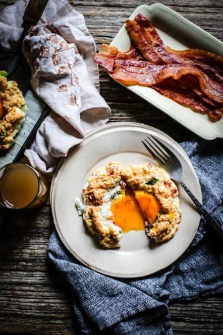 A cloud egg is cut in half on a cream colored plate, there is egg yolk spilling out onto teh plate. The plate is on a black and white napkin on top if a weathered wooden table. there is a small plate of bacon and a glass of juice on the table too.