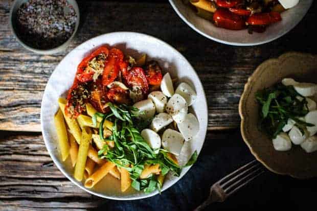 A white bowl is filled with pasta, oven roasted tomatoes, basil ribbons, and mozzarella balls. The bowl is on a weathered wooden table top along with a small bowl of mozzarella;la and basil and a shallow dish of ground pepper.