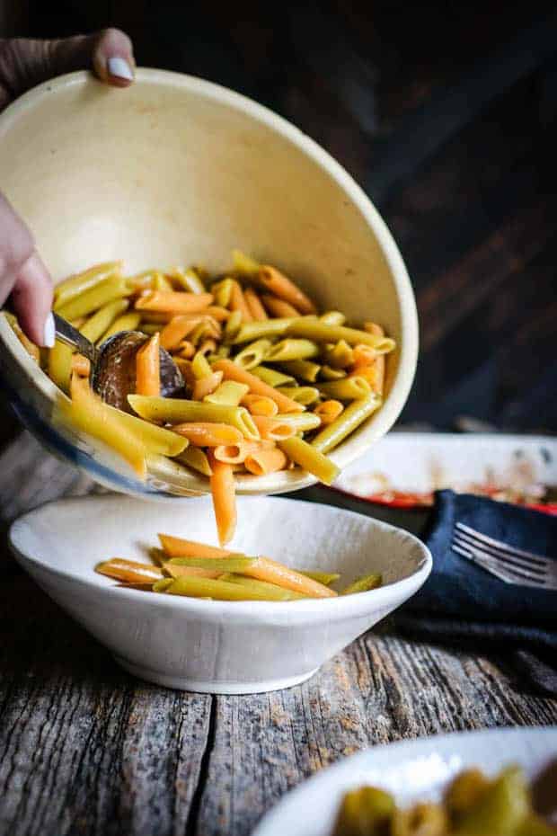 Red and green penne pasta is being poured from a large cream colored mixing bowl into a smaller white serving bowl. The serving bowl is sitting on top of a weathered wooden table top. 