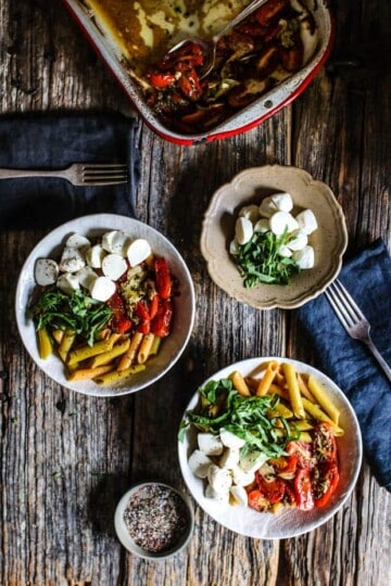 2 white bowls filled with pasta are topped with roasted tomatoes, mozzarella balls, and basil ribbons. The bowls are on a wooden table top with black linen napkins and metal forks. There is a small handmade bowl filled with more balls of mozzarella and basil leaves. At the back of the table there is a white enamel roasting pan with red edges that has roasted tomatoes in it.