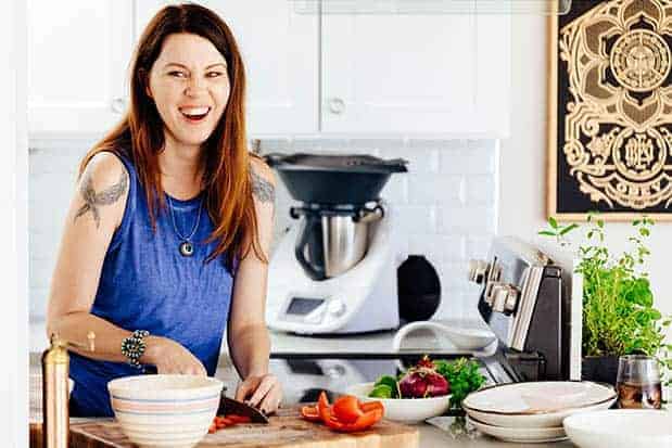 A woman in a blue shirt with long brown hair is in a white kitchen chopping vegetables on a cutting board. There are serving dishes on the counter in front of her. 