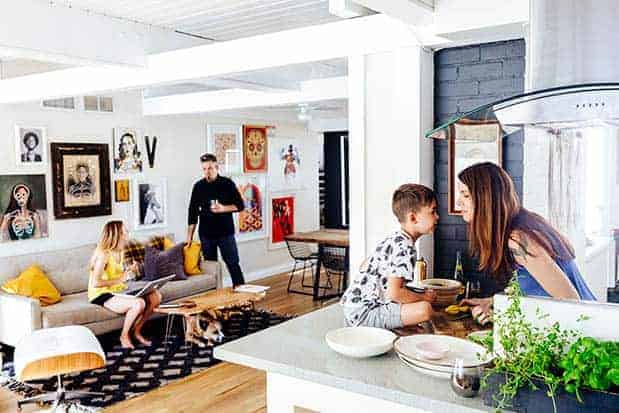 The kitchen and den of a modern home. The walls are white an black with lots of colorful paintings hanging on them. There is a man standing and talking to a teenage girl who is seated on the couch while in the kitchen a mother is leaning over the kitchen counter to give her young son a kiss.