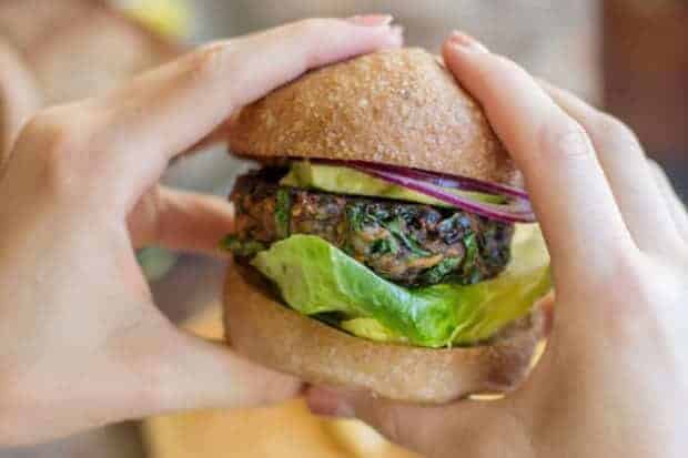 A woman hands are holding an autumn spiced veggie birger , it has the burger patty which is laced with shreds of kale and sweet potato, for garnishes on the burger there is a large piece of butter lettuce, thinly sliced red onion, and slices of avocado
