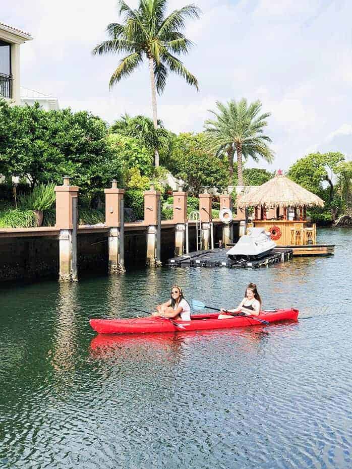 2 girls in a red canoe on the water in Boca Raton Florida. There is a floating tiki hut and palm trees behind them. 