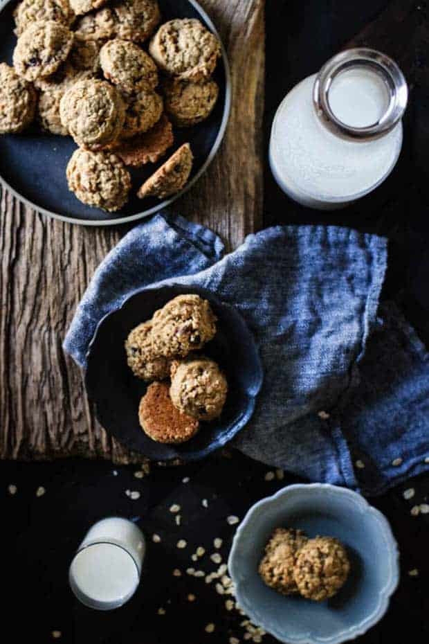 3 bowls piled high with oatmeal date cookies are sitting on a wooden table. There is a large glass jug and a small glass full of milk and a linen napkin also on the table. 