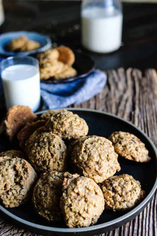 A pile of Gluten Free Oatmeal Date Cookies on a black plate. There is a small glass and a large glass bottle of milk another small bowl filled with cookies and a linen napkin in the background.