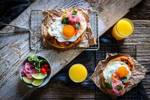 A wooden table top is set with a breakfast scene. 2 glasses of orange juice, 2 small, personal, wire baking wracks with handles each have a large slice of hummus toast on top. There is also a small bowl of garnishes; lime wedges, red pickled peppers, pickled red onions, cilantro leaves. 