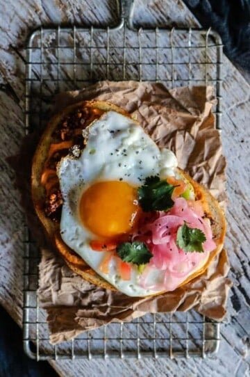 A large, thick piece of toasted sourdough is topped with hummus, walnut taco crumbles, a fried egg, pico de gallo, and pickled red onions.