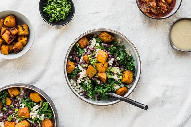 2 large bowls and 3 small bowls on a white table cloth. The large bowls are filled with adzuki beans, miso-glazed kabocha squash, rice, and kale. The small bowls are each filled with extra ingredients, roasted squash cubes, scallions, kimchi, and tahini dressing