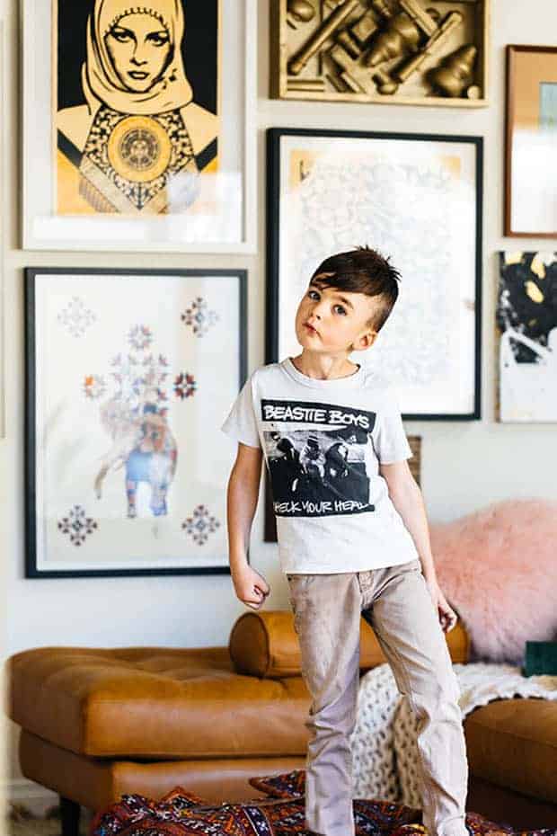 a cute little brown haired boy in tan skinny jeans and a black and white Beastie Boys t-shirt stands in front of a wall full of framed street art.