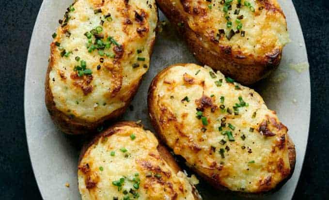 4 twice baked potatoes are sitting on a gray, oval serving platter. The potatoes are topped with melted white cheddar cheese and chives.