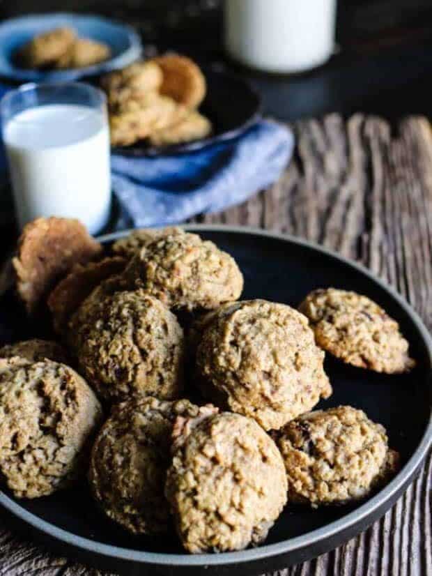 A pile of Gluten Free Oatmeal Date Cookies on a black plate. There is a small glass and a large glass bottle of milk another small bowl filled with cookies and a linen napkin in the background.