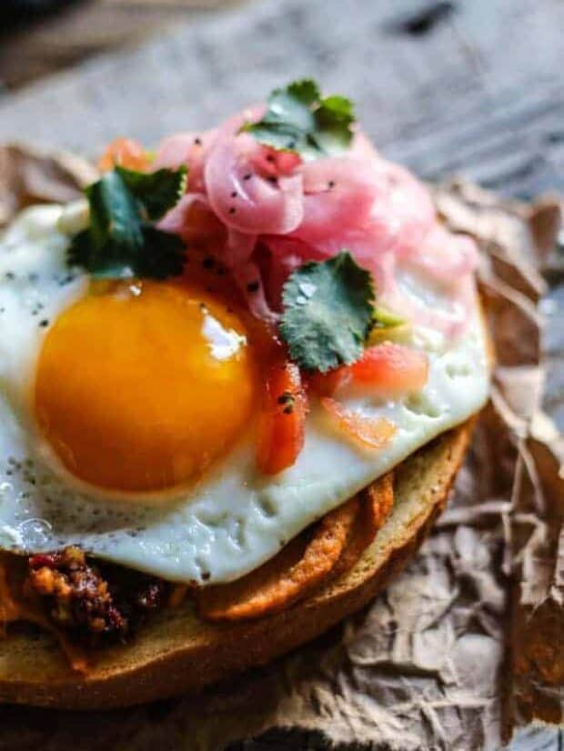 A close up image of hummus toast on a piece of brown butcher paper. On top of the slice of toasted sourdough is hummus, walnut taco crumbles, a perfectly fried egg, pico de gallo, cilantro, and pickled red onions