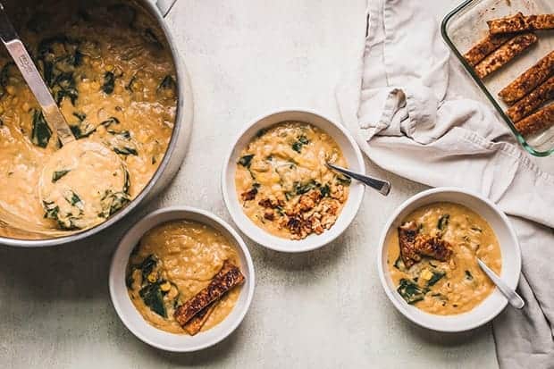 A large pot and 3 small bowls filled with yellow split pea chowder. The small bowls are topped with slices of tempeh bacon. 