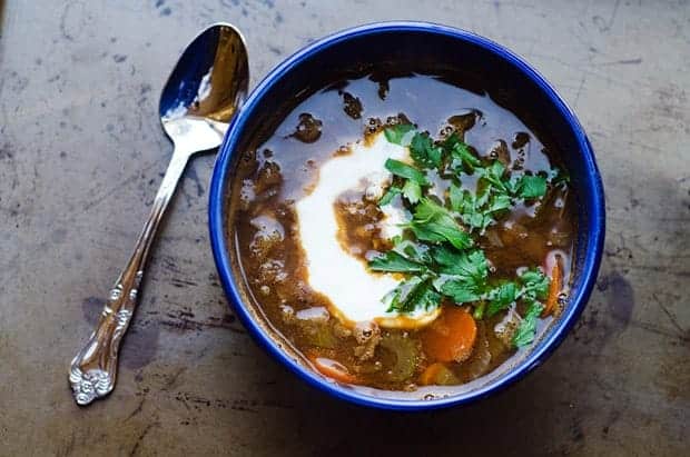 A blue bowl of lentil soup is on a table top and has a metal spoon sitting next to it. The soup is garnished with a swirl of yogurt and chopped parsley 
