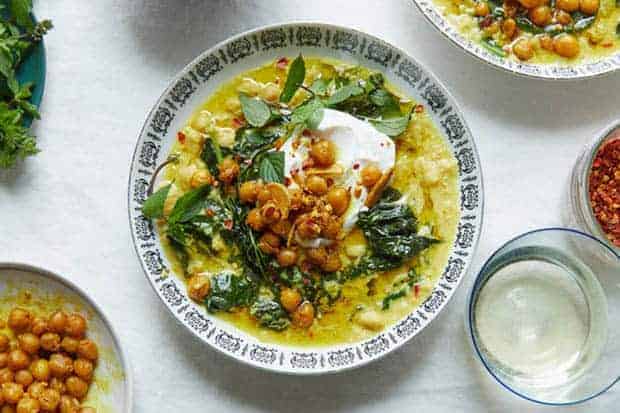 A white bowl with a black design around the edges is filled with Spiced Chickpea Stew With Coconut and Turmeric. There is also a small bowl of spiced chickpeas on the table and fresh mint. 