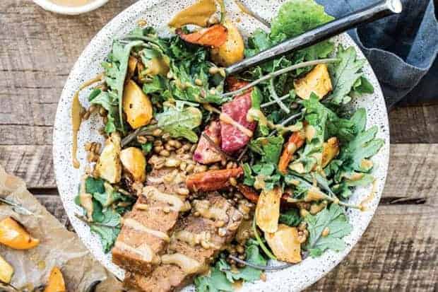 A white bowl filled with greens, tempeh bacon strips, roasted sweet potatoes, and drizzle din a maple mustard dressing