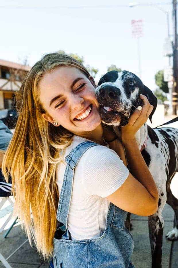 Girl in a blue jean overall dress with a white t-shirt underneath and blonde hair is hugging a black and white spotted great dane.