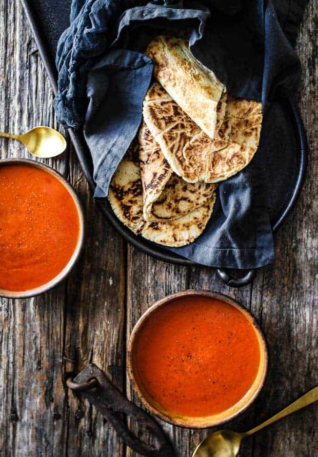 2 bowls of tomato soup on a wooden table top with a basket of grain free naan wrapped in a black linen towel