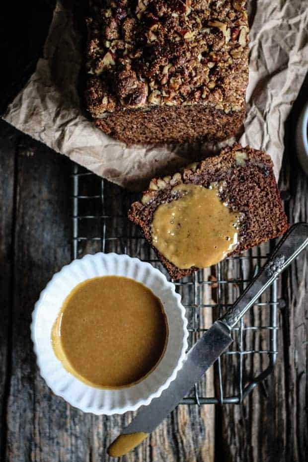 A loaf cake and a slice of loaf cake set on a baking rack. There is a butter knife and a small bowl of maple butter. The slice of loaf cake has maple butter spread over it.