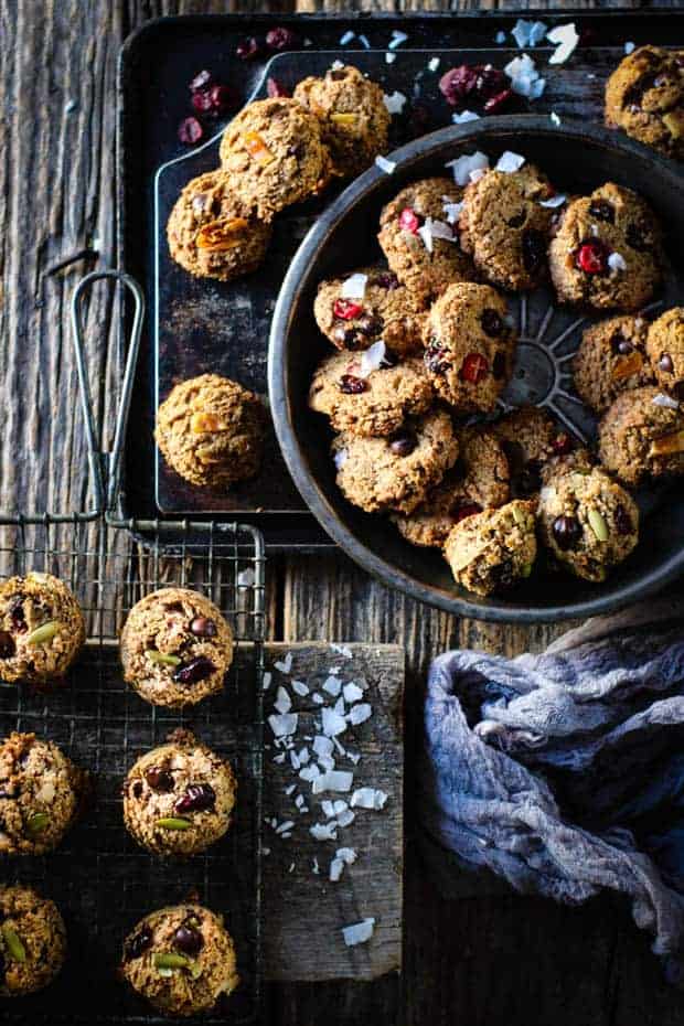 3 different types of kitchen sink cookies arranged in metal bowls an pans on a table top. . There are also coconut flakes and craisins scattered across the tops of the cookies.