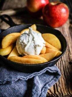 A small skillet filled with vanilla scented fried apples and a scoop of vanilla ice cream. The skillet is sitting on top of a herringbone pattern napkin. There are 2 red apples in the background.