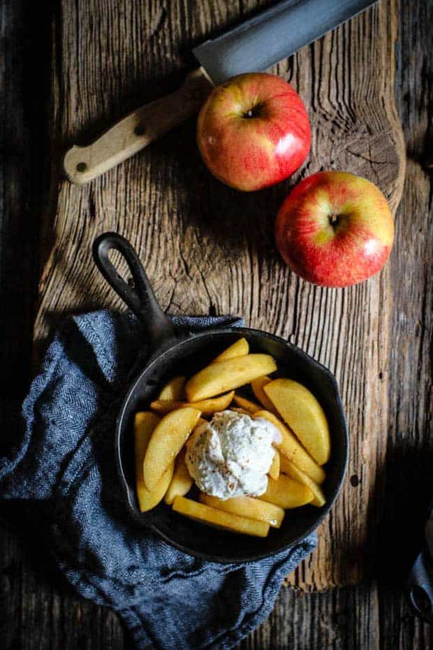 a small cast iron skillet filled with vanilla scented fried apples and a large scoop of vanilla bean ice cream. There are 2 red apples on the table next to the skillet and teh skillet is on a herringbone napkin.