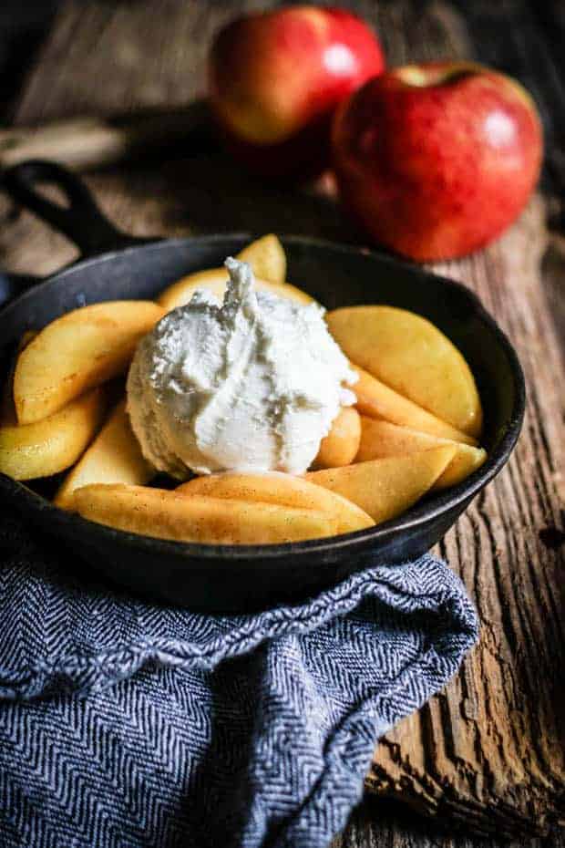 A small skillet filled with vanilla scented fried apples and a scoop of vanilla ice cream. The skillet is sitting on top of a herringbone pattern napkin. There are 2 red apples in the background.