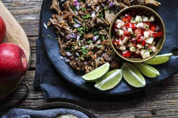 Instant Pot Carnitas garnished with chopped red onion and cilantro on a black plate with lime wedges and a bowl of apple salsa sit on a wooden table that also has a cutting board with whole apples on it.