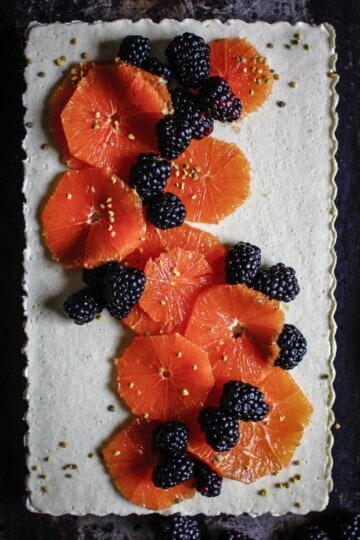 A white tart topped with slices of orange and blackberries with a sprinkle of bee pollen.