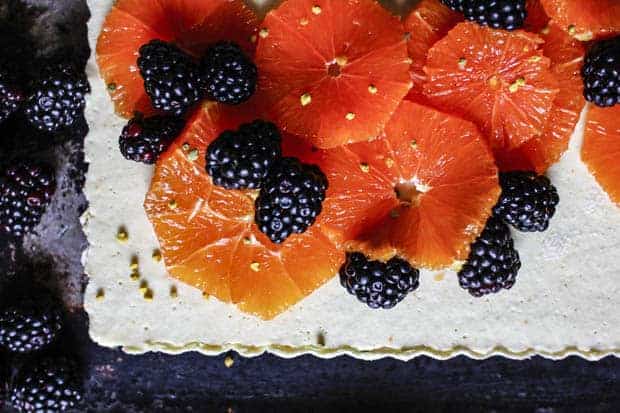 A horizontal image of a white tart with fluted edges topped with sliced, pink, Cara Cara oranges, blackberries, and a sprinkle of yellow bee pollen.