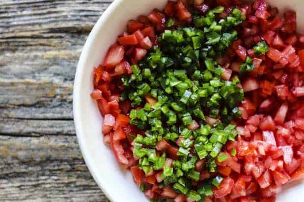 A shallow white bowl is filled with seeded and finely diced tomatoes and minced jalapeños. Teh bowl sits on a weathered wooden table top. 