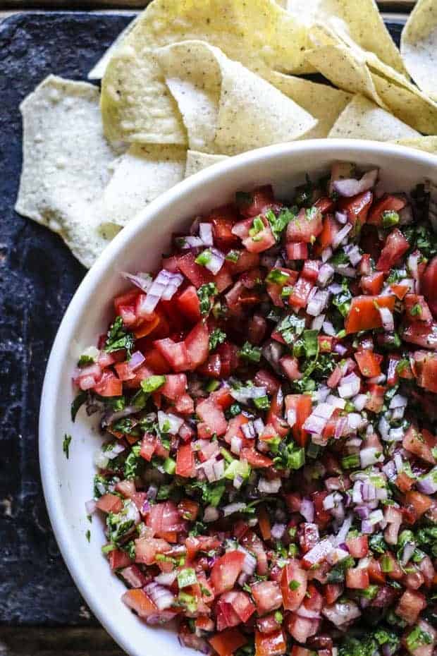 A white bowl filled with pico de gallo. Finely diced tomatoes, red onion, cilantro, jalapeño, and citrus juice. There are tortilla chips around the bowl.