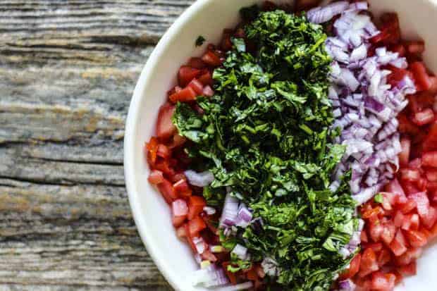 A shallow white bowl is filled with seeded and finely diced tomatoes, minced jalapeño, red onion, and cilantro. The bowl sits on a weathered wooden table top. 