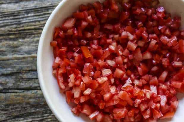 A shallow white bowl is filled with seeded and finely diced tomatoes. Teh bowl sits on a weathered wooden table top. 