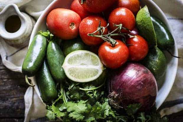 A large, white shallow bowl is filled with red Roma and on the vine tomatoes, jalapeño peppers, a red onion, limes and lime halves. The bowl is sitting on a white linen napkin with blue stitching and there is a small ceramic pitcher next to the bowl. 