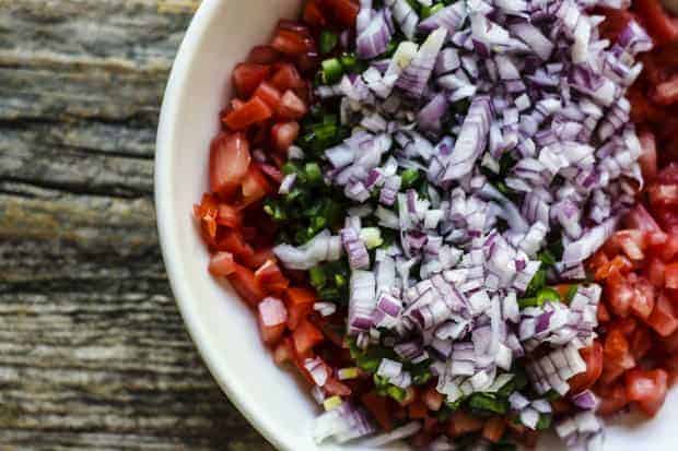A shallow white bowl is filled with seeded and finely diced tomatoes, minced jalapeño, and red onion. The bowl sits on a weathered wooden table top. 