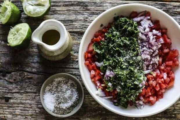 A shallow, white bowl filled with pico de gallo ingredients - layers pf finely diced tomatoes, red onion, jalapeño pepper, and cilantro. A small dish of coarse salt, a small ceramic pitcher of olive oil, and 3 squeezed lime halves are ob the table next to the bowl. 