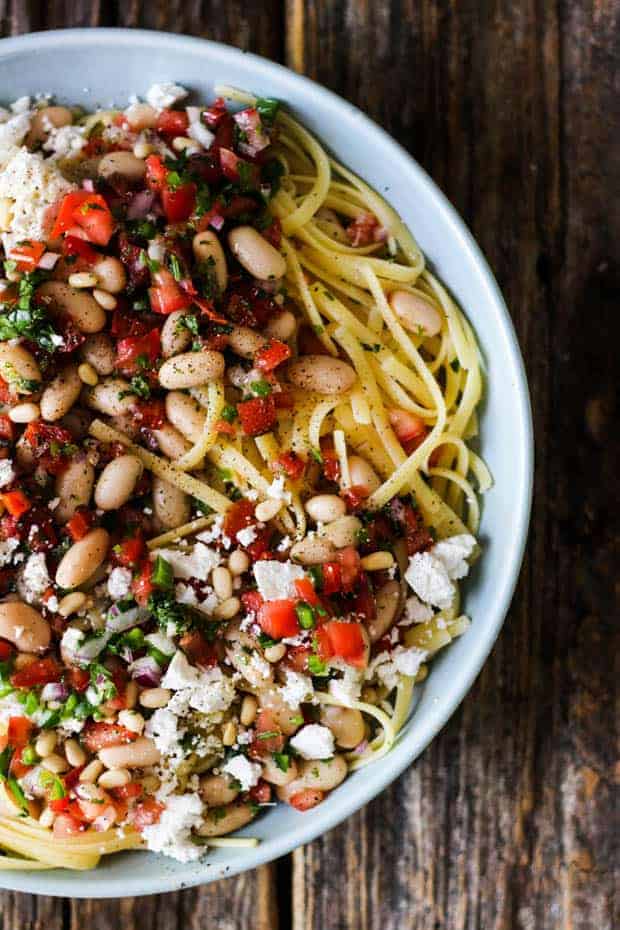 A shallow light blue bowl is on a wooden table, filled with fettuccini pasta, white beans, pico de gallo, and crumbled ricotta salata cheese.